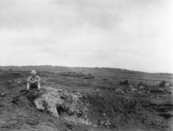 View of a soldier seated on rocky ground looking off to the left. Behind him and to the right a row of soldiers are behind sandbags, and behind them are a row of tanks. On the far right is a jeep, a tent and a cart.