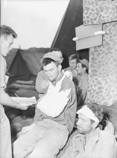Marine corporal with his arm in a sling after being shot in the upper arm at Iwo Jima. Another wounded soldier sits beside him with a bandage on his head.