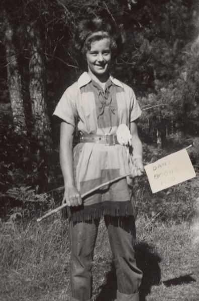Sue Ann Hackett standing outdoors in her first-prize Daniel Boone costume, along with badge prize attached to her belt. Sue Ann is holding a sign on a stick that reads, "Daniel Boone 1950."
