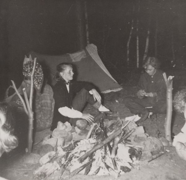 Margo Pirie and Sue Ann Hackett on an outdoor camping trip. They are seated near a campfire with a tent in the background.