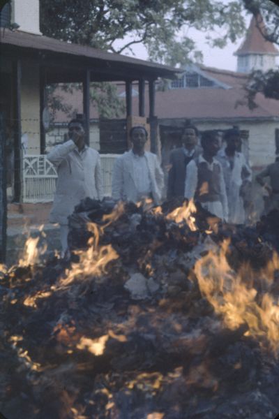 Bank burning money in Tezpur, Assam during the Sino-Indian Border Conflict. The large pile of paper is steadily burning and a group of men watch from behind. There are buildings in the background.