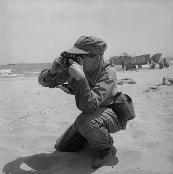 Dickey Chapelle taking photographs on the beach in Milwaukee during Operation Inland Seas. A Navy ship is on Lake Michigan in the background on the left. Along the shoreline are marines with another military ship.