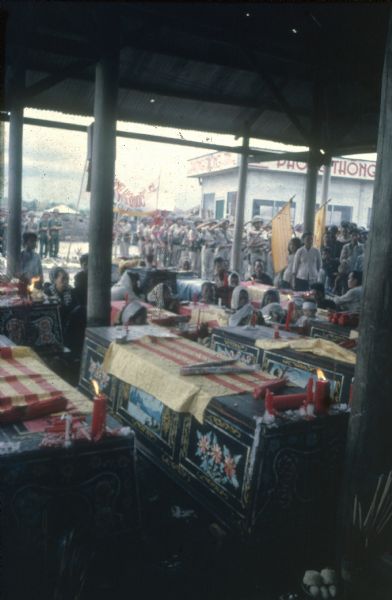 Funeral at Binh Hung, Vietnam for several members of the Vietnamese Sea Swallows battallion who were killed in combat against Viet Cong forces on May 29. Under a roofed building with columns are elaborately decorated caskets draped with flags, with candles burning at the end of each one. Mourners are sitting among the caskets, and lines of soldiers paying their respects are lined up outdoors in the background.