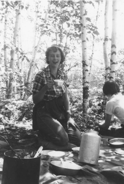 Sue Ann Hackett kneeling and holding a tin cup on an outdoor camping trip. There is a tarp in the foreground with dishes and food containers. Birch trees are in the background. Another camper sits with her back to the camera at right.
