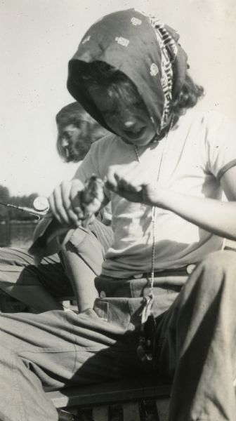 Close-up of a young girl wearing a bandana on her head sitting in a boat and removing a fish she has caught from the hook. Another girl is behind her holding a fishing rod.