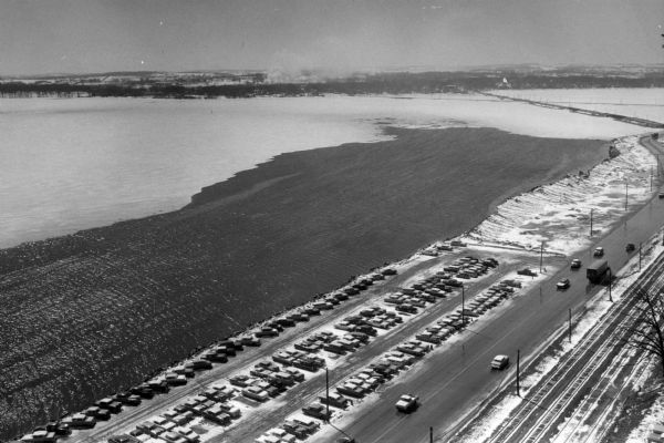 Elevated view of a parking lot along John Nolen Drive on the shore of a partially frozen Lake Monona. Railroad tracks are in the foreground on the right.
