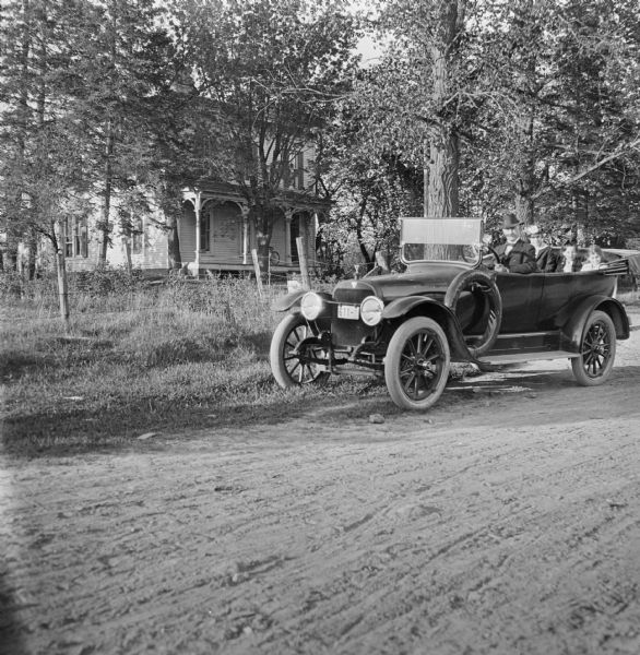 Family of four seated in a car near the Wisconsin State Horticultural Society orchard. The car is parked on a country road in front of a house.
