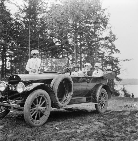Three women sit in the back of a Hudson Touring car, while a fourth woman stands behind the car on the side rail. All four women wear scarves over their hats. The car is parked in front of trees near a lake. Two men stand near the shoreline in the background.