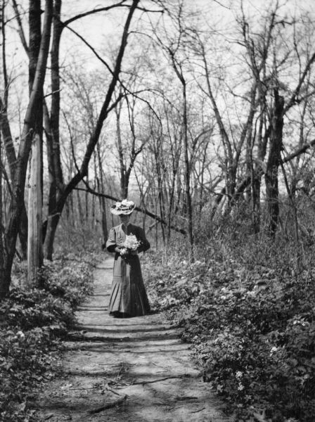 Mary E. Smith walking down a sunlit path through trees. She is carrying a bouquet of freshly picked flowers.
