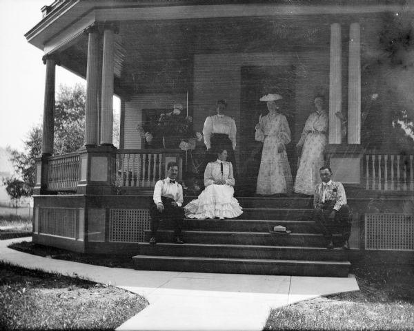 View from walk of several members of Dr. Joseph Smith's family grouped together on a porch. Mary E. Smith is standing in the center.