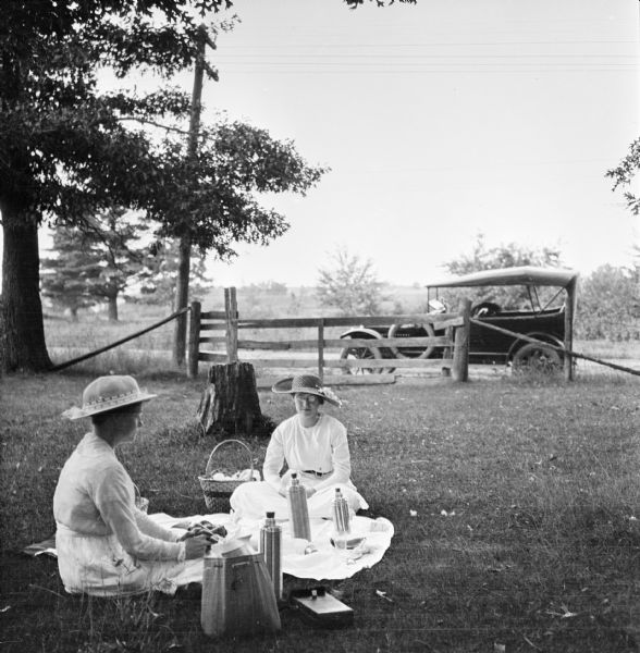 Roadside picnic on the way to Waupaca. Mary E. Smith is seated with a friend on a white blanket with thermoses and a picnic basket. In the background the automobile is parked on the side of the road next to a fence.