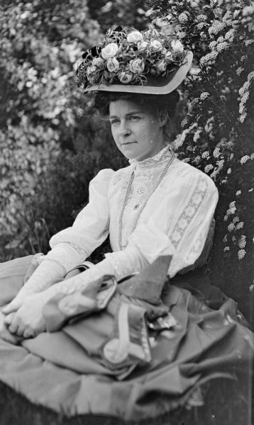 Portrait of Mary E. Smith sitting outdoors on the grass in front of a flower bush. She is wearing an ornate hat decorated with roses.