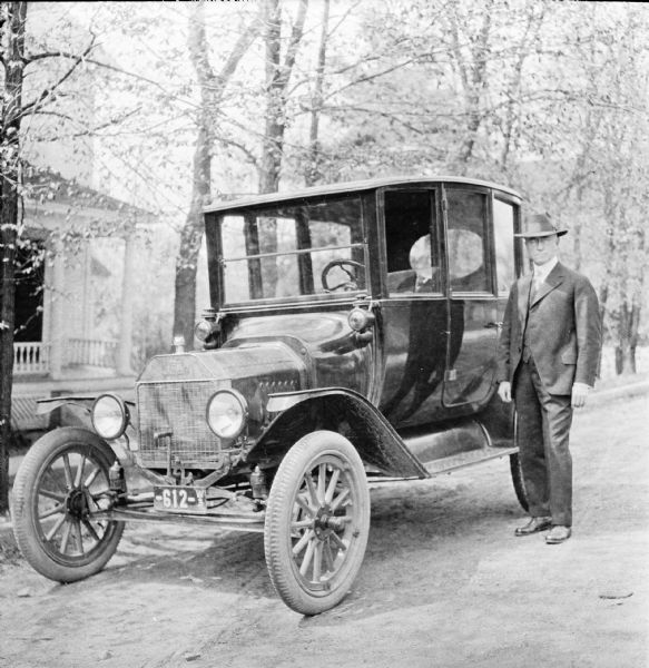 Dr. Joseph Smith standing next to his Ford automobile. The automobile is parked along the curb of a tree-lined street in front of a house.