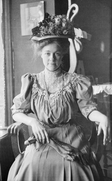 Indoor portrait of Mary E. Smith seated in a chair. She is wearing an ornate hat topped with roses and is holding a draped pair of black gloves in her lap.