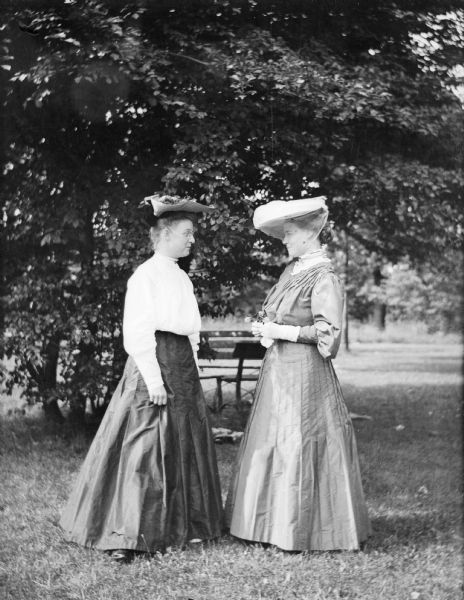 Mary E. Smith with an unidentified women standing in a park near a bench. Mary is holding several flowers in her hand.