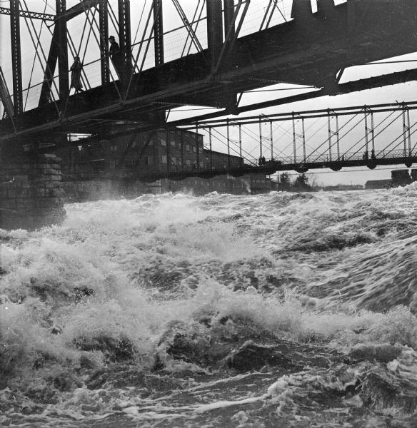 Water of Big Bull Falls or the Wisconsin River rapidly flows underneath two railroad bridges.  Pedestrians cross the bridges while peering over at the overflowing river below.