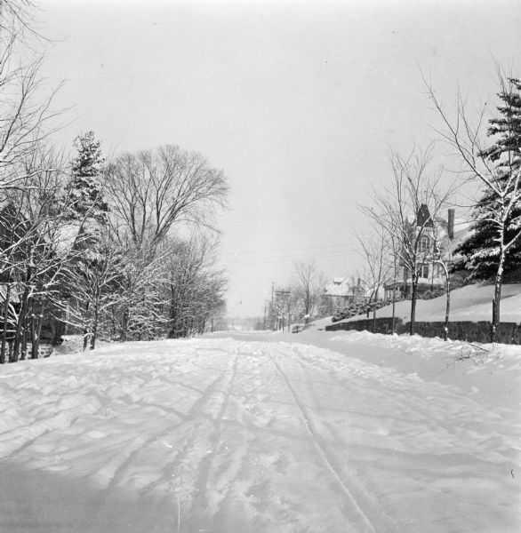 Winter scene with a road covered with freshly fallen snow. Houses and trees line either side of the road and are also covered in fresh snow. Vehicle tracks cut through the snow on the road.