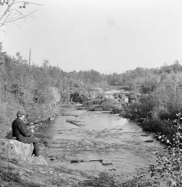 Elevated view of man seated on cliffs edge overlooking the dells of the Eau Claire River. Farther upstream is a bridge over the river.