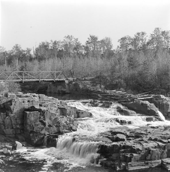 Elevated view of the Eau Claire River rushing over large rocky formations. A bridge crosses over the river on the left. Large tree trunks have been stacked along the far side of the river.