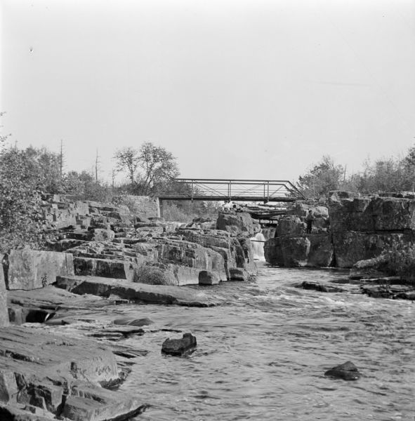 View up river towards a bridge over a rocky portion of the Eau Claire River. A women is standing on rocks beneath the bridge in the center, while a man stands on a large rock near the riverbank on the right.