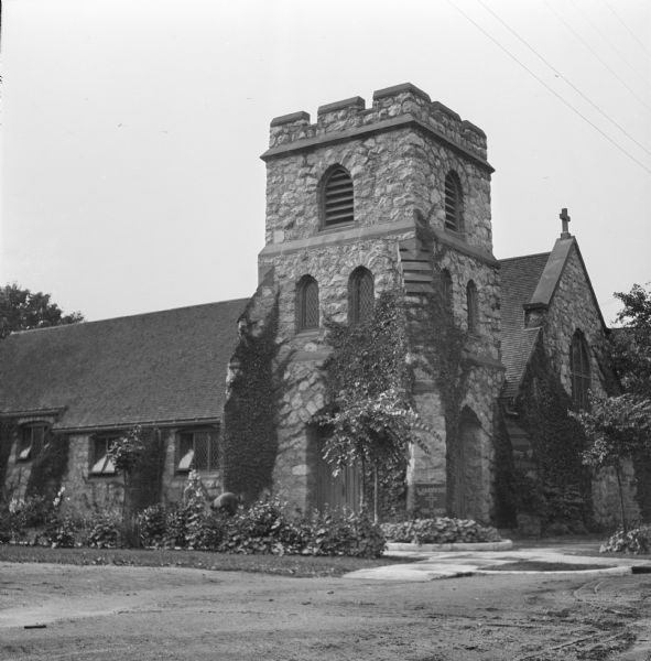 View of side and front of the St. Ambrose Episcopal Church. Two people are standing near the ivy which is growing up the side of the tower near the arched door behind a strip of plants and flowers.