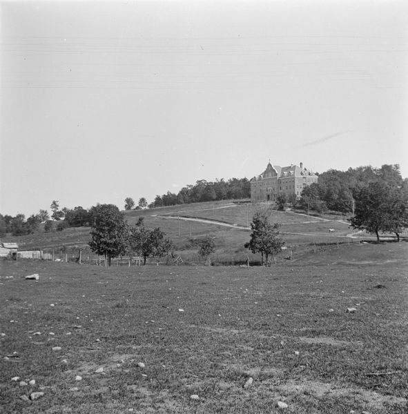 View from field looking up at St. Mary's Hospital situated atop a hill in front of fenced pastures. A horse is grazing in a pasture on the right.