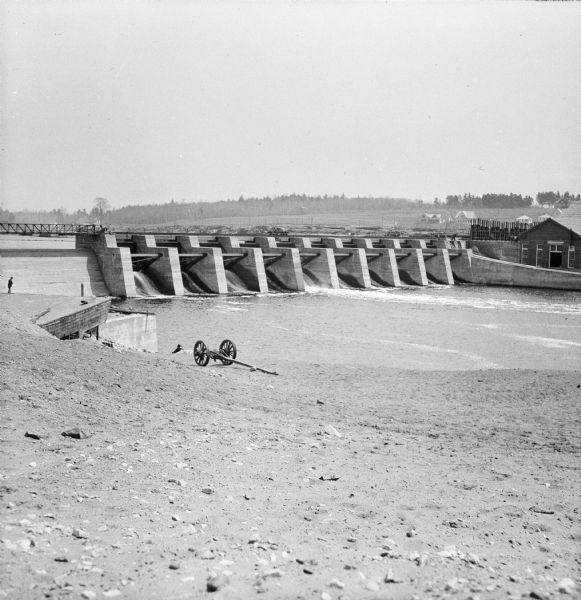View down riverbank towards dam of a river around Wausau. Two men and a limber (two-wheeled cart) are near the rivers edge. A man, perhaps a construction worker, stands on the dam near a set of ladders. Several houses are on a hill on the opposite side of the river.