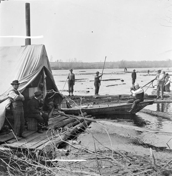 Businessmen visiting a log drive down the Wisconsin River. One man wearing a vest sits in a boat holding a peavey hook over his shoulder. Other men wearing shirts and neckties stand on logs and a boat to the right of him. On the left is the cooks tent with three men aboard. Other men are standing on top of logs further out in the river using pike poles to move them. There are buildings along the hilly riverbank in the far background.