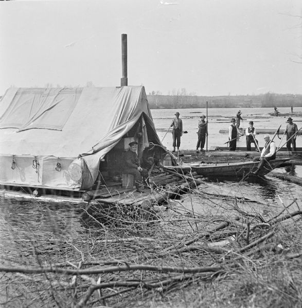 View from shoreline over tree branches of three businessmen visiting a logging crew on the Wisconsin River. One businessman sits on a boat while two others pose in a boat behind him. All three man are holding peavey hooks and wearing vests and neckties. Several log drivers stand on logs and hold pike poles used to maneuver the logs down the river. On the left two men sit on the raft which hold the cooks tent which is tied to the shoreline. There are buildings along the hilly riverbank in the far background.