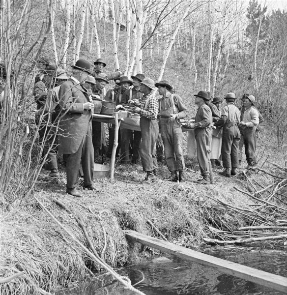 View towards shoreline of log drivers eating lunch on the edge of the riverbank. The men wait in line along a buffet table filled with provisions. The businessmen are wearing suits, dress shoes, hats and ties. The rest of the men are wearing work clothes and work shoes. A steep hill with birch trees rises behind them.