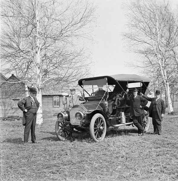 Four businessmen in suits and hats stand around an automobile parked among birch trees, while another man sits in the passenger seat. They are visiting a log drive. Wood buildings are in the background.