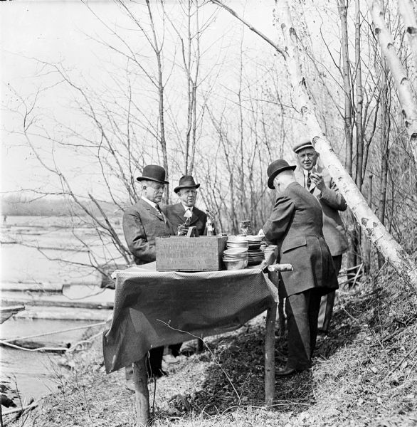 Four businessmen standing around a table, eating along the banks of the Wisconsin River. The table is set with a stack of bowls and a crate of apples. Logs and log drivers float along the river.