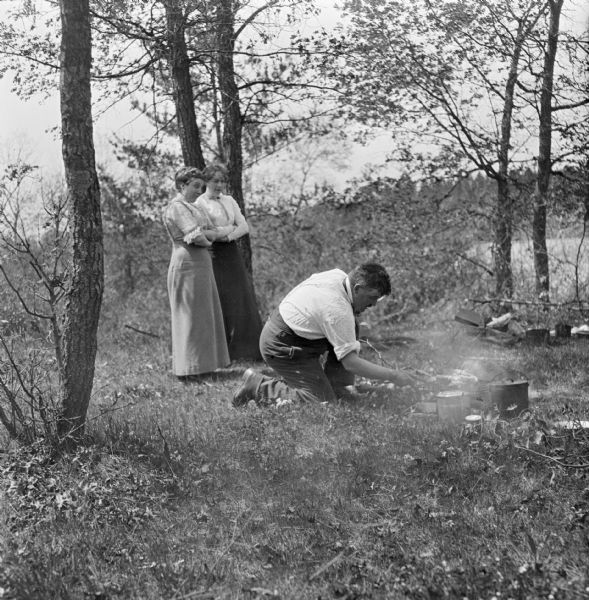 Marvin B. Rosenberry cooks over a campfire in a wooded area, while Katherine Landfair Rosenberry and Mary E. Smith stand behind him watching.