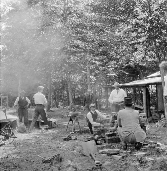 Rotary Club barbeque along the Plover River at the campsite of Neal Brown. Several men gather around a barbeque pit where meat is being cooked on a rotisserie.