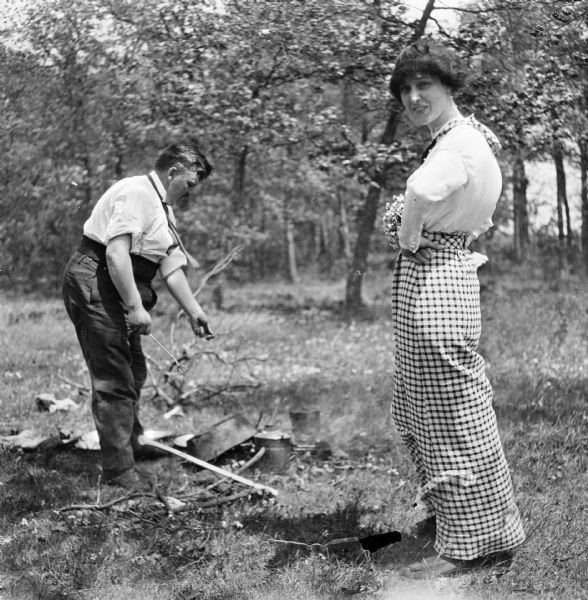 Marvin B. Rosenberry cooking over a campfire while a young woman, possibly his daughter (?), holding a bouquet of flowers watches.
