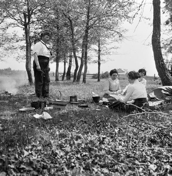 Mary E. Smith, Katherine Landfair Rosenberry, and possibly Marvin Rosenberry's daughter (?) sit in the grass having a picnic. Marvin Rosenberry stands near a campfire overlooking the women.