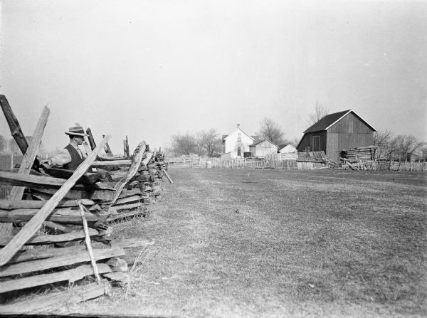 Man leaning against a split-rail fence. A farmhouse and several farm buildings stand along the far parameter of the fence.
