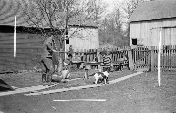 A man pumping a water pump that drains into a water bucket behind a fence. Cows behind the fence are drinking from the bucket.  Next to the man is a young boy petting a dog.