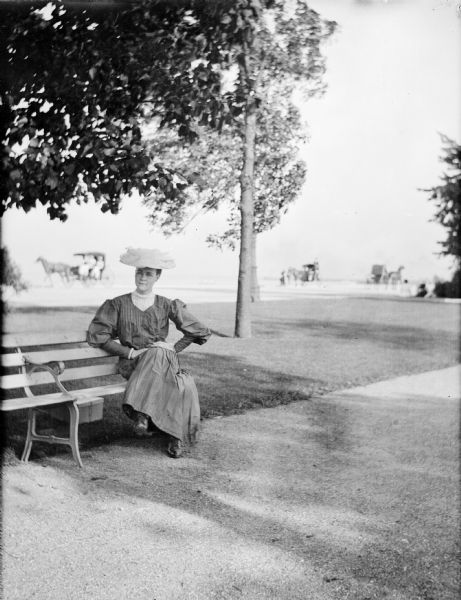 Mary E. Smith posing on a bench in Jackson Park. Several horse-drawn carriages are on the street behind her.