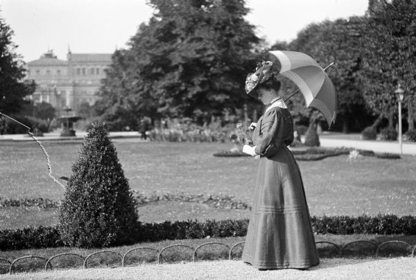 Mary E. Smith walking along a path in the Volksgarten in Vienna, Austria. She is wearing an ornate hat, holding an umbrella over her shoulder, and looking down at the rose that she is holding in her gloved left hand.