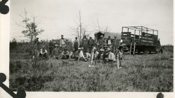 Group of men posing in a field. There are two trucks parked behind the group, and axes are propped up in the foreground.