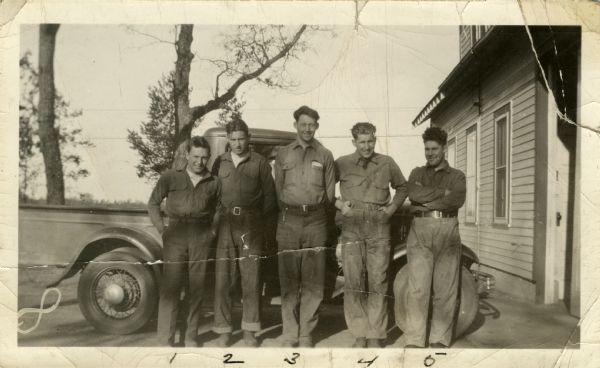 Five men from the Civilian Conservation Corps Camp Petenwell stand together and smile for the camera. They are posed in front of a truck which is parked next to a wood frame building on the right.