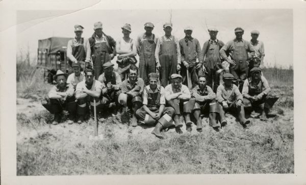 Group of men posing in two rows in a field. The men sitting and crouching in the front row are wearing folded down rubber boots. The men standing behind them are wearing overalls. There is a truck parked in the background.