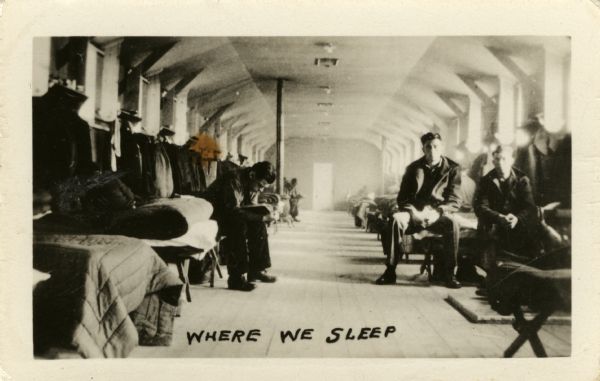 View down a long room with beds lined up in rows along the sides for Civilian Conservation Corps members at Camp Petenwell. There is a woodburning stove in the background on the left. A few men sit on their beds.