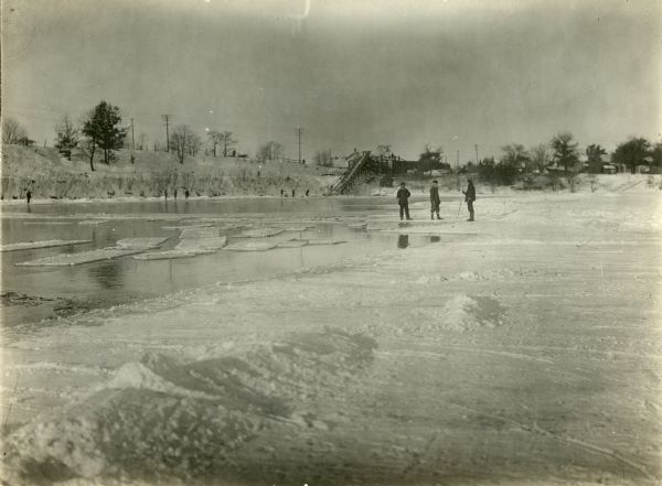 View from ice of men from the CCC group at Camp Petenwell cutting ice. Three men stand on the ice on the right. A steep bank surrounds the icy water. Men are along the far bank, and more men are standing near the shoreline. In the background is a wood chute leading up to an industrial building where more men are standing. There is a horse standing on the bank near the chute.