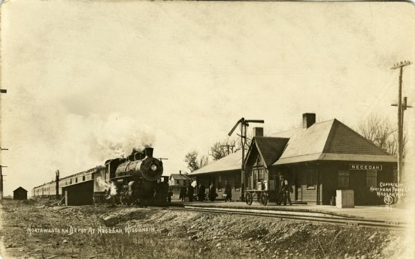 View from side of railroad tracks of a train at the depot. A few men stand on the platform, and a man is pulling a luggage cart.