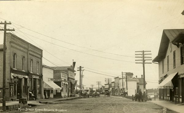 View down unpaved street towards many businesses: Carl Olson Shoe Store, Wakeman's Pharmacy, Jay Jennings, dentist, O.G.H. Parman, Badger Hardware, L.G. Bishop Meat Market, J.E. Daly Drug Store, J. Williams & Bros. Hardware, Carl Olson Shoe Store, a restaurant, the post office, and Jacob Gross General Store. There are multiple horse and wagon vehicles parked or rolling down the street. Men are standing, sitting and walking along the street. A unicycle is parked outside the general store.