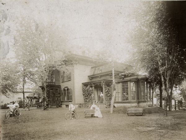 An Italianate style home, with a small balcony above the front entrance. The windows on the main section of the house are arched and have matching arched shutters, which are open. There are seven people posed outside the home, on the lawn and on the porch. A man and woman stand on a porch in the center, with a dog lying between them. On the lawn are two women posed near a bench, and a man poses perched on a bicycle next to them. On the far left, a woman stands near a young girl who is standing and holding a bicycle.