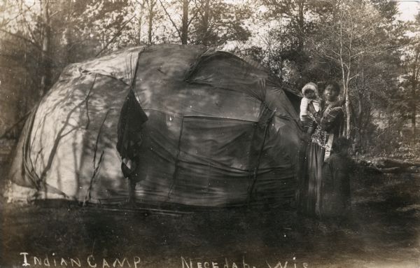 A Native American Ho-Chunk woman stands holding a baby outside a wigwam. Another child stands just in front of the woman, looking off to the right. The photograph has been annotated 'Indian Camp Necedah, Wis'.