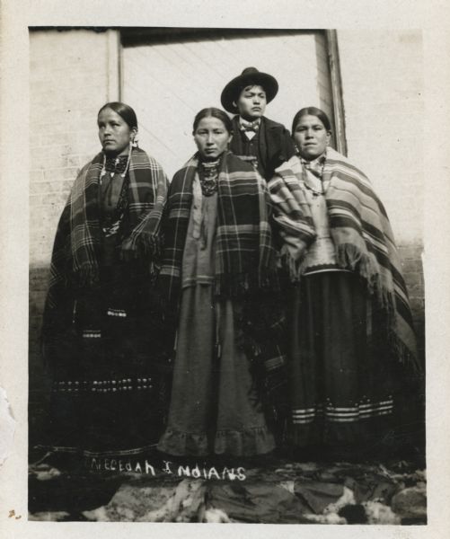 Three women and a young man pose in front of the wood door of a brick building. The man stands behind the three woman, and is wearing a hat, suit coat and bow tie. He appears to be holding a cigar. They are traditionally dressed. The photograph is annotated "Necedah Indians."